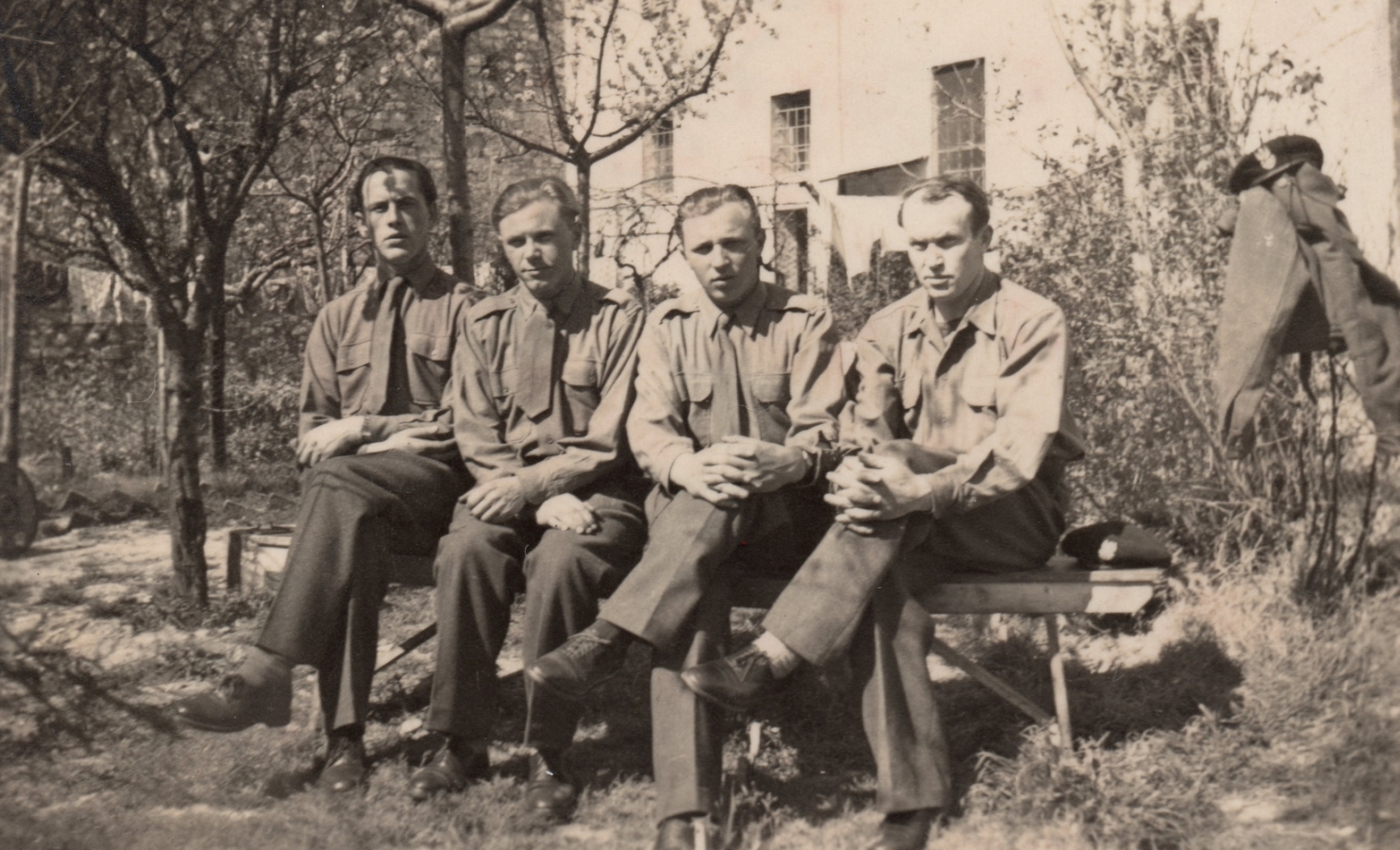 Four soldiers 
sitting on a bench under trees in front of a house, in shirtsleeves, no caps, one cap next to the men on the bench, another 
placed on a jacket hung over a wire fence.