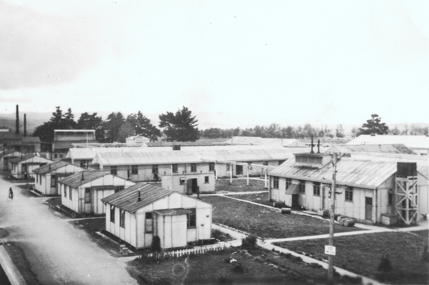 Closer 
shot of the teachers' quarters. Several small prefabricated buildings with their vertical joints clearly visible, sitting 
between a road and patches of lawn.