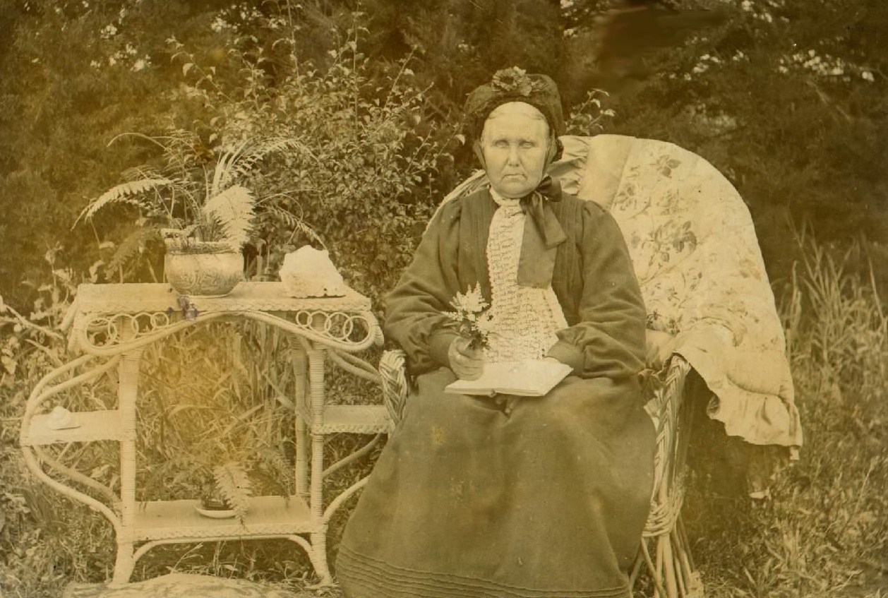 The old lady seated 
on a wicker chair in the garden, next to a wicker table and with a book on her knee.