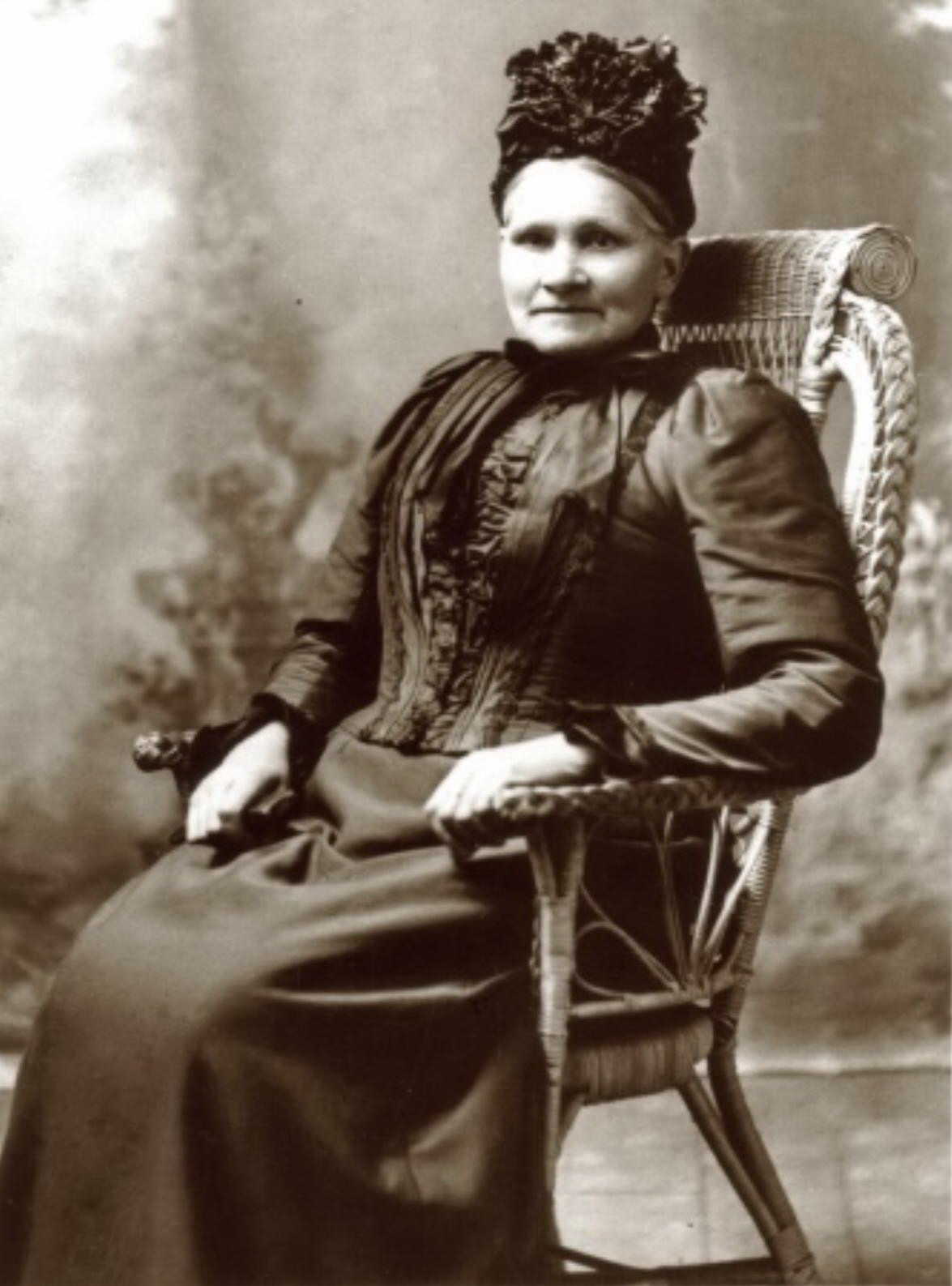 A studio 
photograph of a middle-aged Rosalia in hat and posh dress seated on a wicker chair