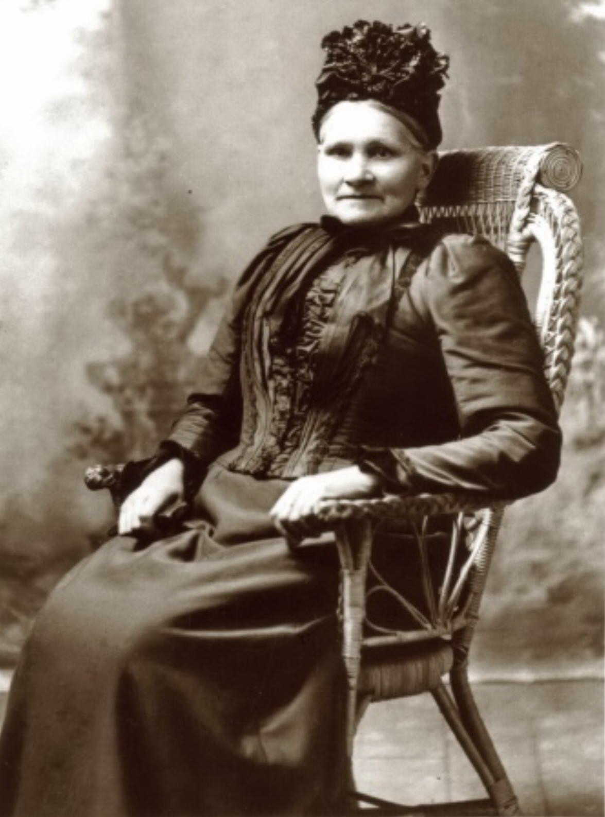 A studio 
photograph of a middle-aged Rosalia in hat and posh dress seated on a wicker chair