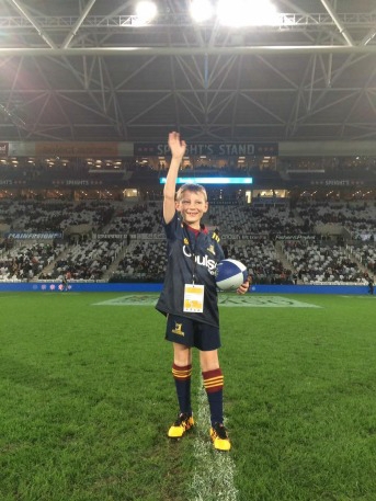 Thomas with his hand 
up and  holding a rugby ball on the centre line of the Forcyth Barr Stadium