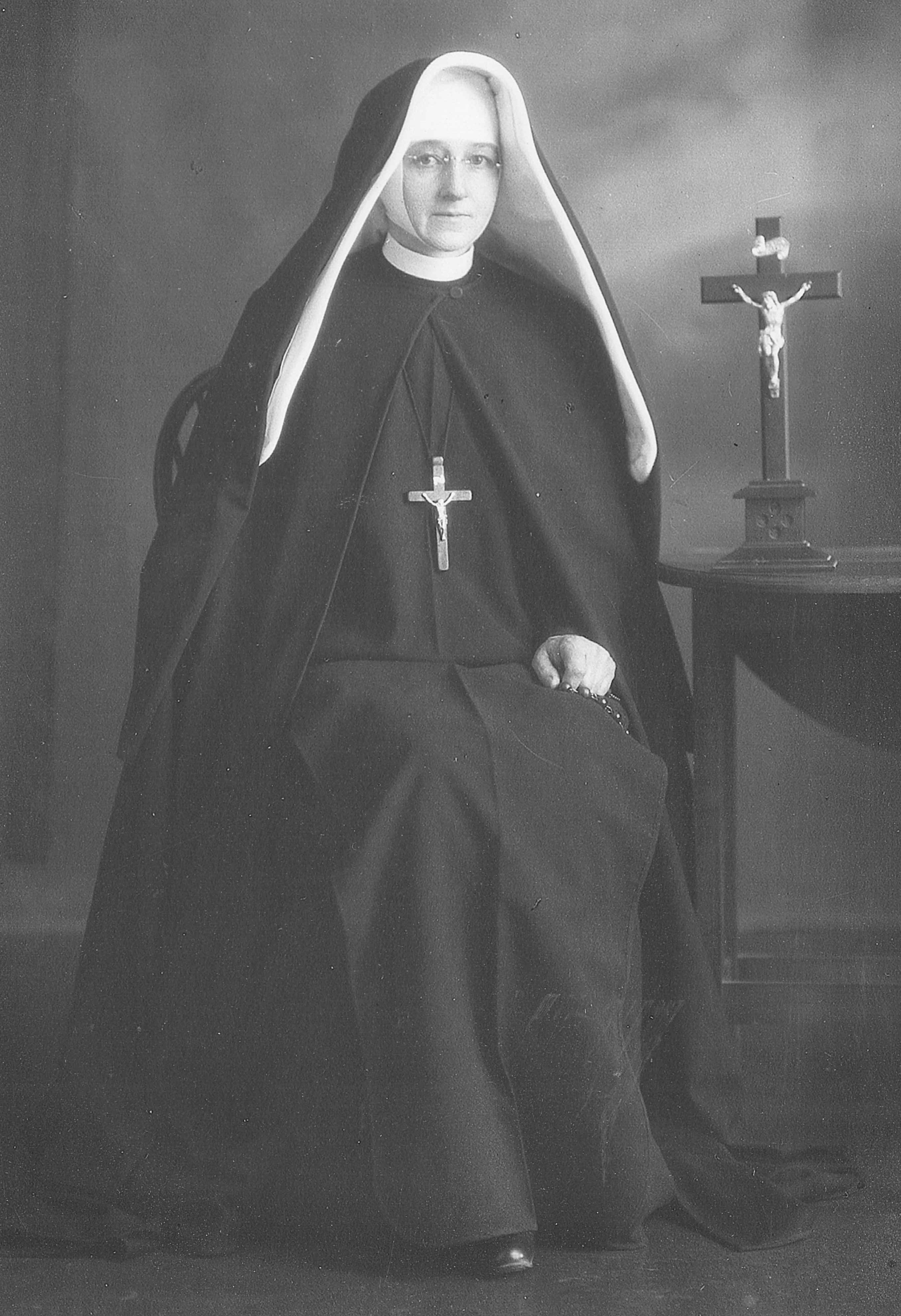 Black 
and white photograph of Sr Mary Kostka in her habit, sitting next to a table with a crucifix on it, and holding her rosary.