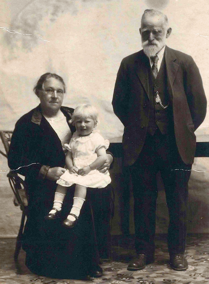 Black and white studio photograph of Benna sitting on Mary Potroz's lap, and with Thomas standing next to them, hands 
behind his back.