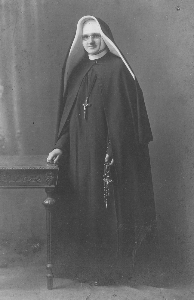 Black 
and white photograph of Sr Dunstan in her habit, standing next to a carved table and holding a large-beaded rosary with a 
crucifix nearly as big as her hand.