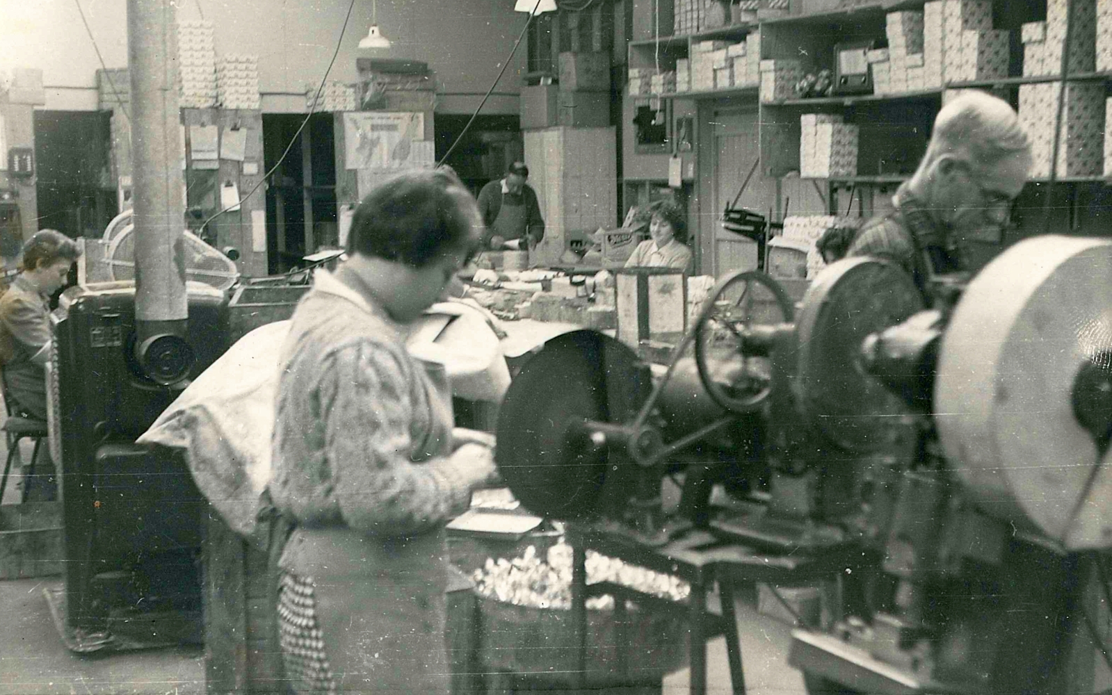 Black and white photograph 
the factory floor. In the foreground is a machine and immediately behind are a young woman and older man working separately. 
In the background are three other people, heads down, at desks, and shelves filled with boxes.