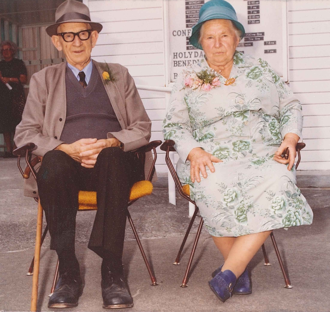 Coloured informal photograph of Jacob and Ellen sitting on chairs with mustard-coloured seats, outside the church. 
Ellen is in a white, long-sleeved dress with green roses and is wearing a turquoise-coloured hat. She looks serious. Jacob, 
in a light-brown hat that matches his jacket and waistcoat, smiles.