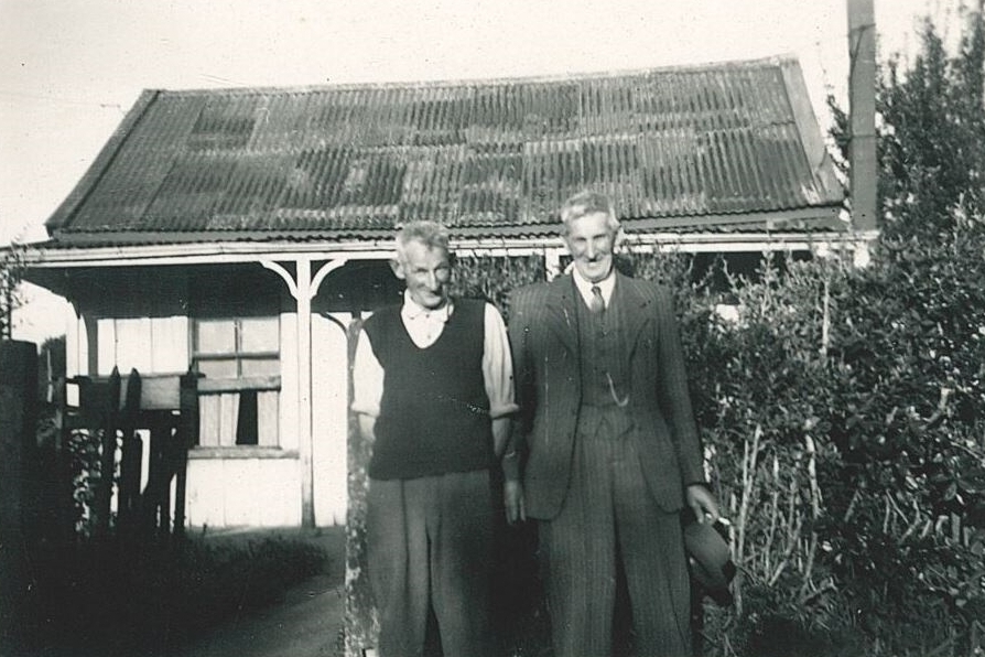 Black and 
white photograph of Jacob and Frank standing outside the old house, Frank in a three-piece suit and holding his hat, and 
Jacob more casual in rolled-up shirt-sleeves and a knitted sleeveless jumper. The dodgy curve in the corrogated iron roof is 
clearly visible.