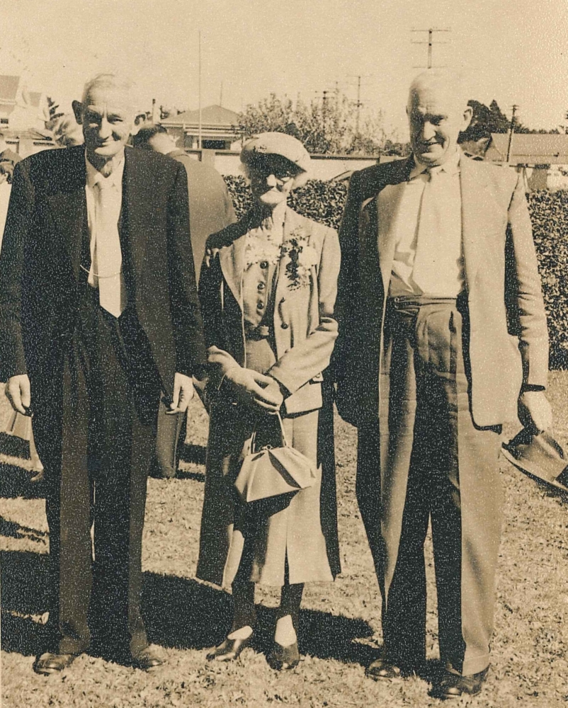 The three standing smartly dressed on short grass and a well-clipped hedge. Frank has a white tie with a watch-chain 
across it, Mary has a hat, handbag and flower corsage on her suit lapel.