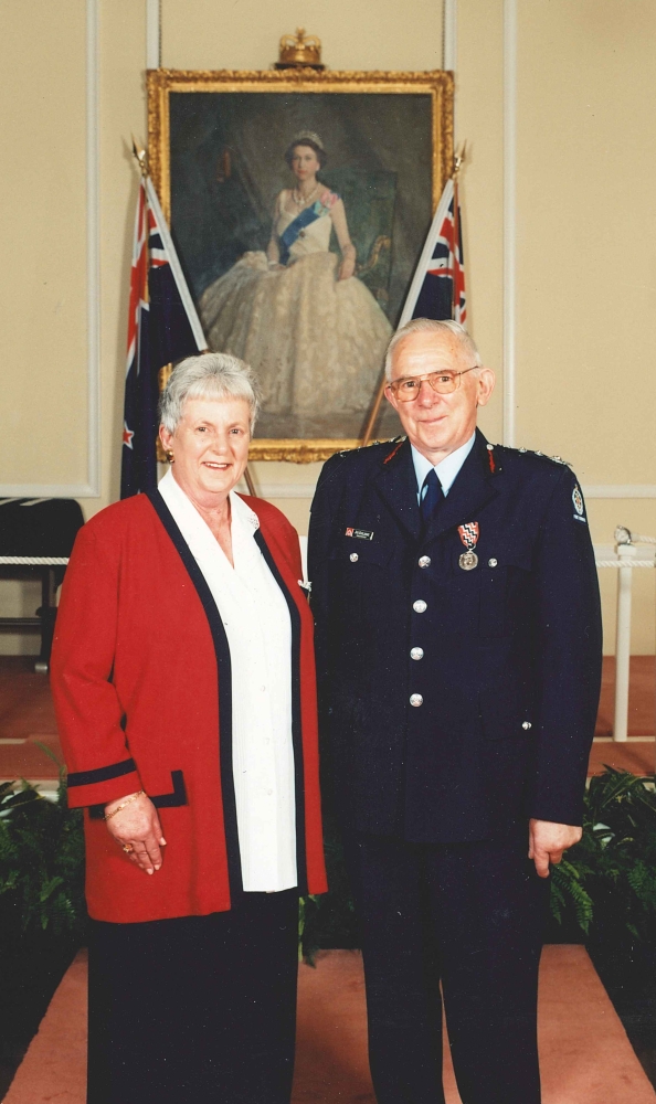 Coloured 
formal photograph of Joe and Patsy Kuklinski in front of a formal portrait of a young Queen Elizabeth II flanked by two New 
Zealand flags. Joe is in his formal fire service uniform, with the QSM. Patsy is in a long black skirt, white shirt and red 
jersey-jacket etched in black.