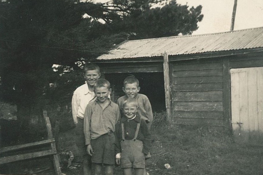 Black and white 
photograph of the four boys, aged about five to , standing close together in front of a rough-hewn single-storey barn, all 
smiling widely, two at the back, two at the front. The one behind the smallest boy is bending slightly and has an 
extra-impish grin.