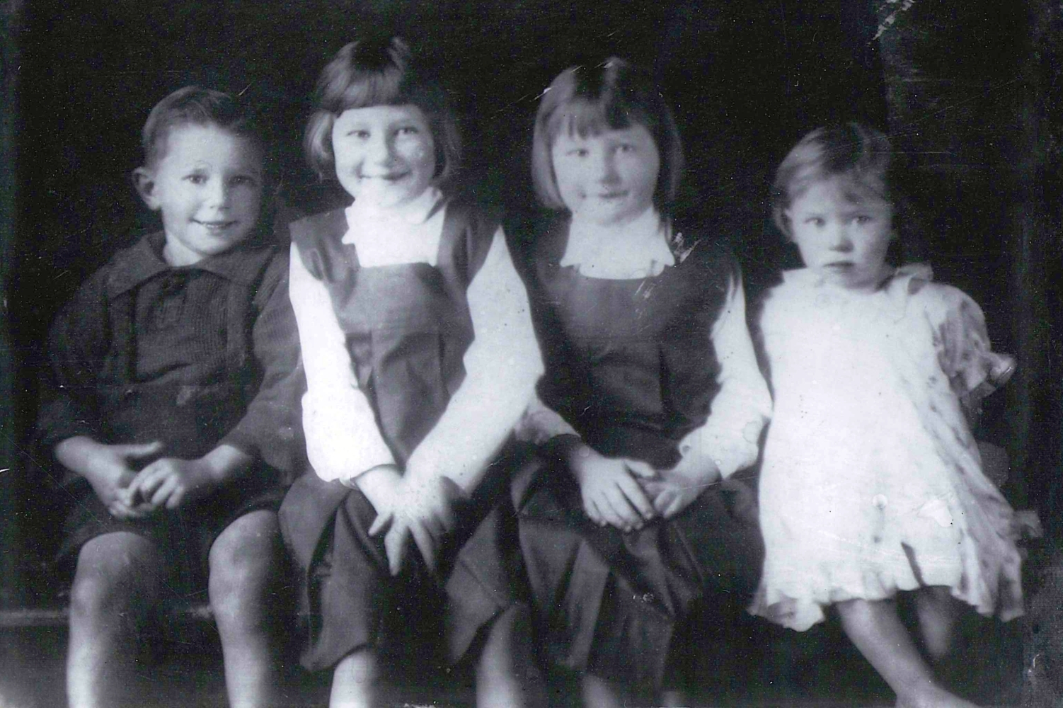 Black and white
photograph of the first four Kuklinski children, sitting: Benna and Pauline in the middle with too-large gym-lips, and with 
similar page-boy hair cuts, Joseph on one side in shorts and a dark shirt smiling as widely as Benna, and Gertie on the other 
side in a frilly dress looking slightly more suspiciously at the photographer.