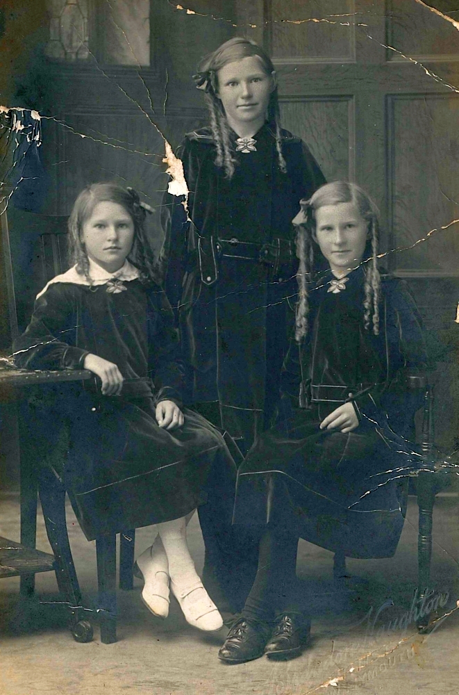 Black and white 
studio photograph of the youngest Potroz siblings in matching dark, possibly velvet, dresses. The three bonde girls all have 
their long, fair hair curled into locks, each with a ribbon in her hair. Ellen and Millie are half-smiling, but the youngest, 
Pat, with her legs crossed at the ankles and not touching the floor, looking more serious.