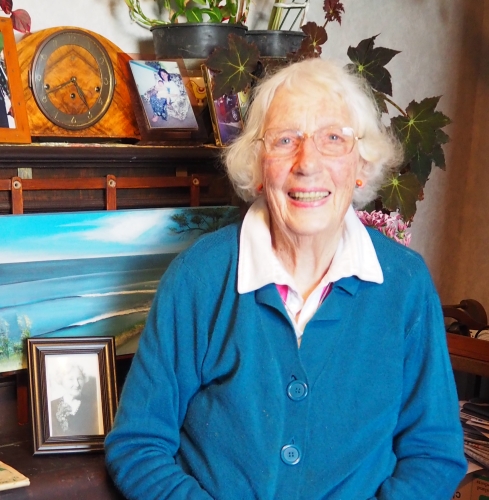 Gladys in a 
blue cardigan sitting at her photograph-covered piano.