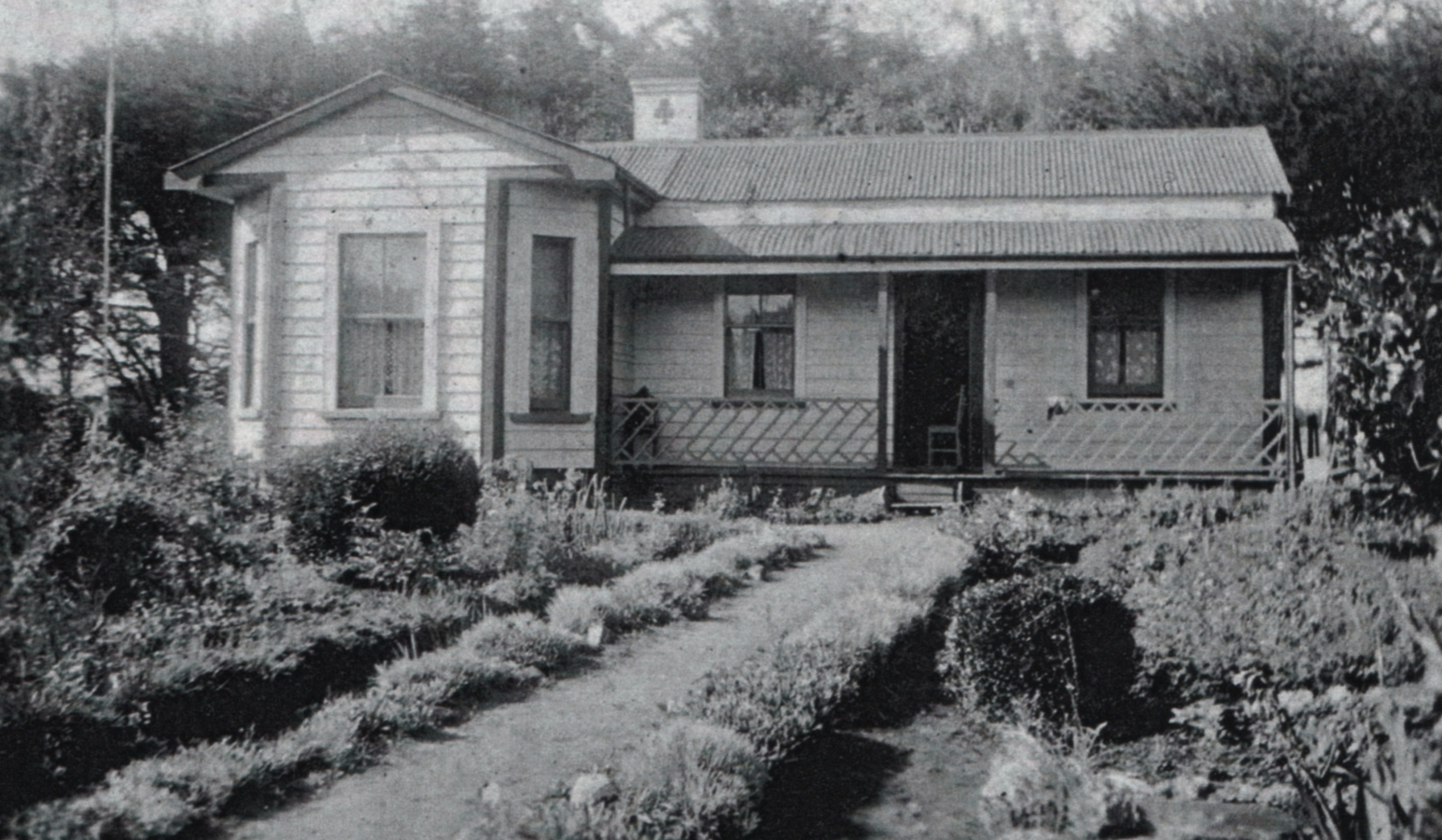 A black and 
white photograph of a bungalow with a neat garden on both sides of a straight path in front.