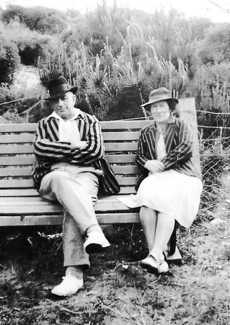 Bert and Edith 
sitting on a bench in front of a fenced hill, both in striped jackets, Bert with arms folded