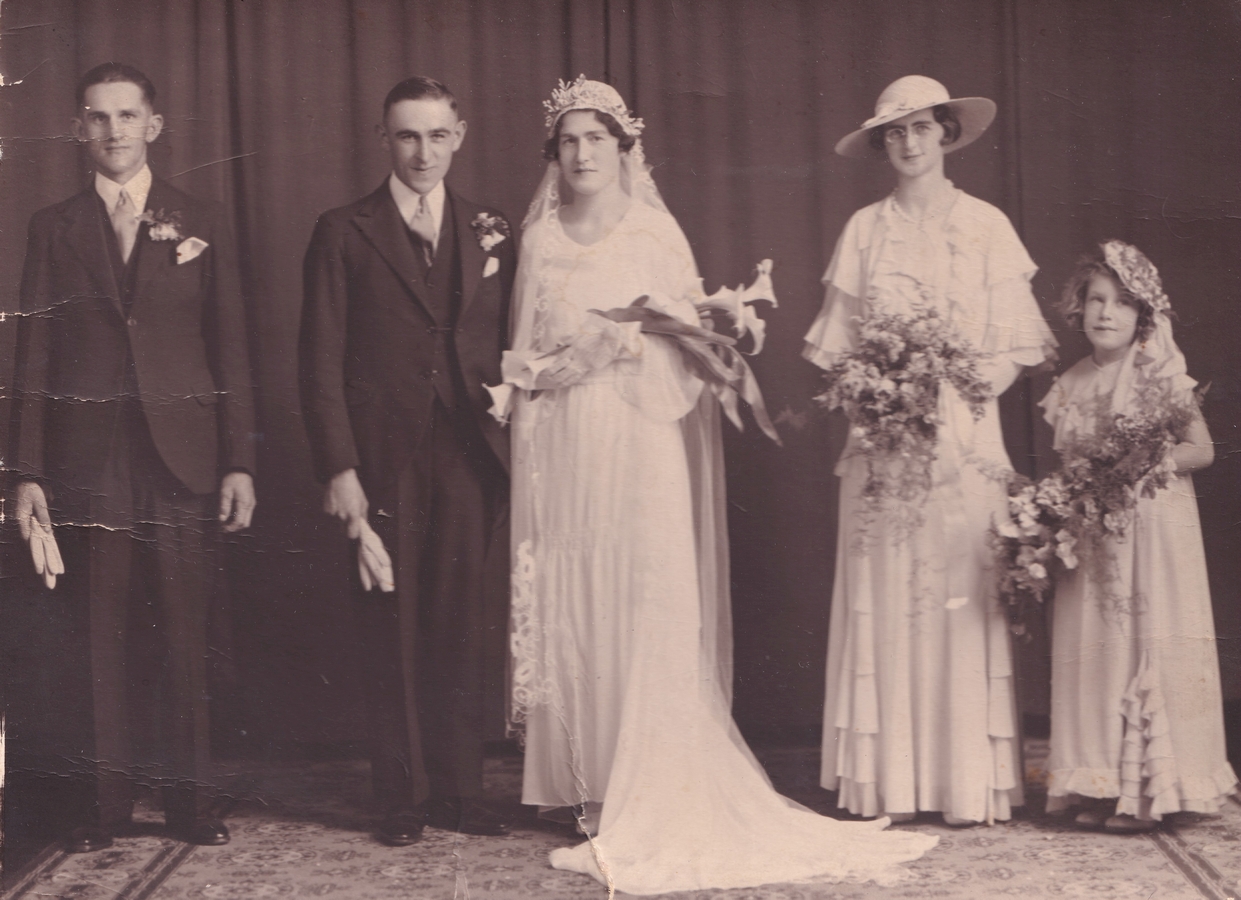 Formal photograph ofwedding party. Joe is standing to the left, both men are holding white cloves, Margaret has a 
   1930s-style cloche head-covering, and the bridesmaid is in a wide hat. Bride, bridesmaid and flowergirl all holding 
   boquets