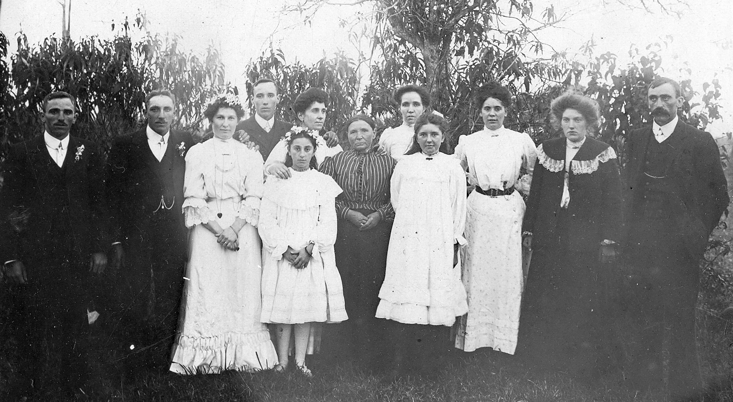 Wedding party of 12, outside. Apolonia has a gentle smile but most look quite serious. Men in dark formal attire and 
most women in light dresses.