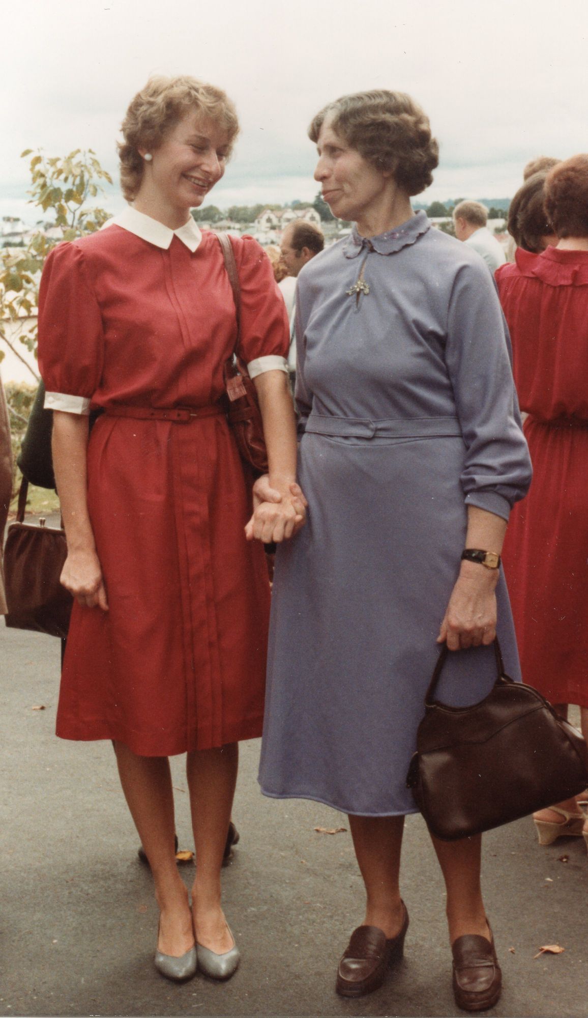 A coloured 
photograph of Ciocia Julia and Regina standing, holding hands and looking at each other and smiling, both wearing similar, 
high-collared dresses. Ciocia Julia's dress is a duck-grey, and she has a handbag and sensible shoes, and Regina's dress is 
red with white trimmings. Her shoes are silver.