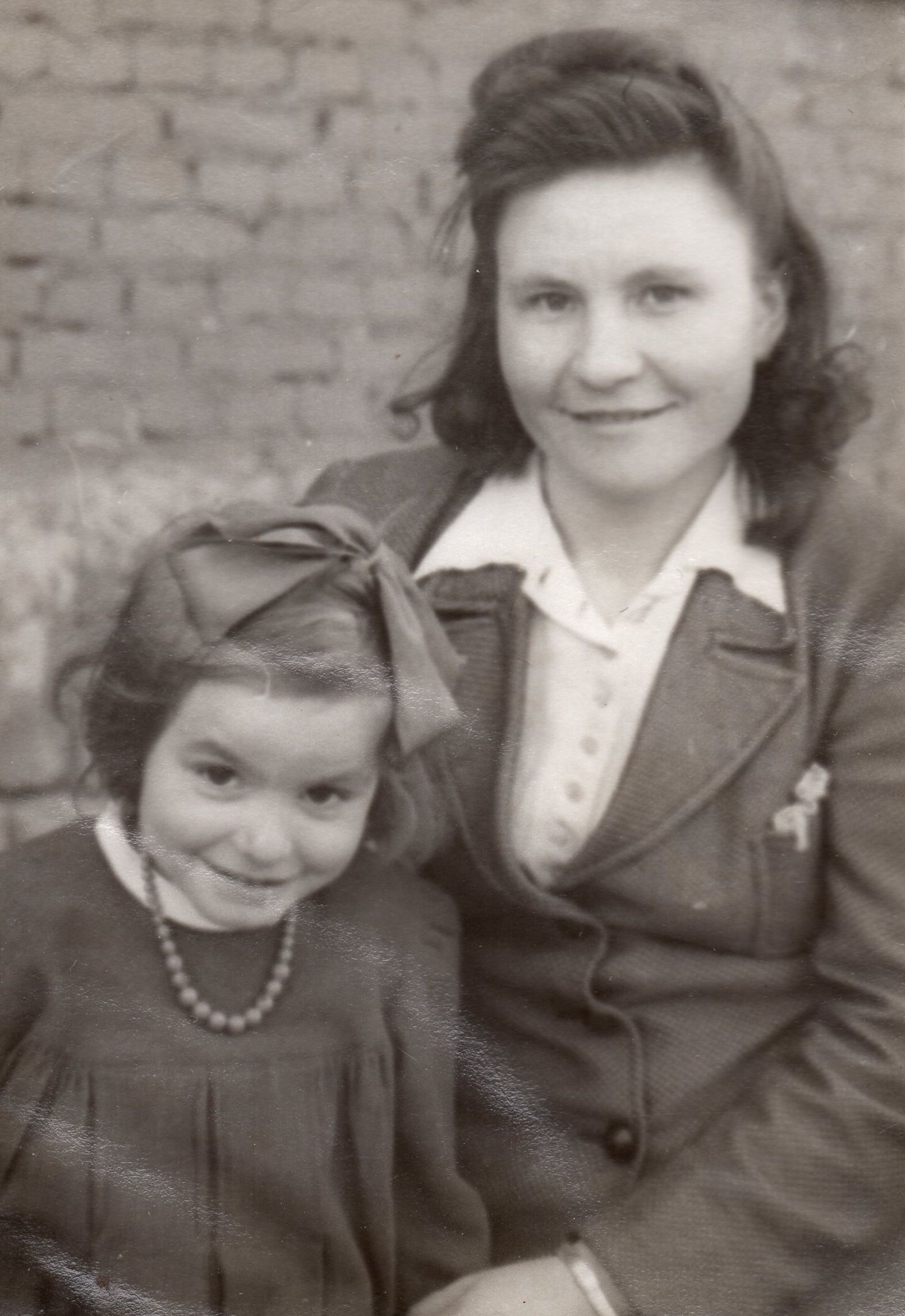 Black 
and white pic taken outdoors. Emilija is seated and wearing a jacket and white shirt and Zenona is wearing a dress pleated on 
a high bodice, with a necklace and a ribbon in her hair. Both are smiling.