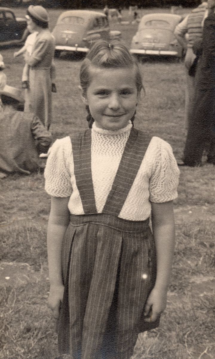 Black 
and white pic of Zenona in a paddock, smiling and wearing a dark skirt and pale short-sleeved crocheted top. In the 
background are parked cars, circa 1950s, and several members of the public doing their own thing.