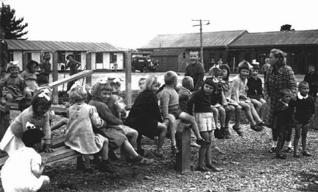 Young children sit along a  
long see-saw. Buildings and soldiers in background and.