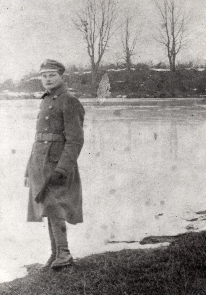 Wojciech Pleciak 
standing on the edge of a lake in thick army coat, looks like snow, bare branches on trees