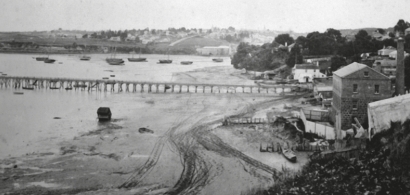 a black and white 
   view of a harbour with boats below hills with houses and a wooden wharf across. Tracks have been made across the sand from 
   the middle foreground towards the wharf