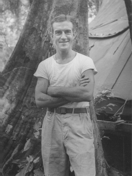 a casual black and white photograph taken beside a tree and with a tent in the background, of Frank in tee-shirt, 
   arms crossed and looking at the camera, smiling
