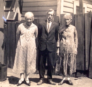 a black and white photograph of three elderly people standing in a back garden looking serious. William is between 
   his sisters
