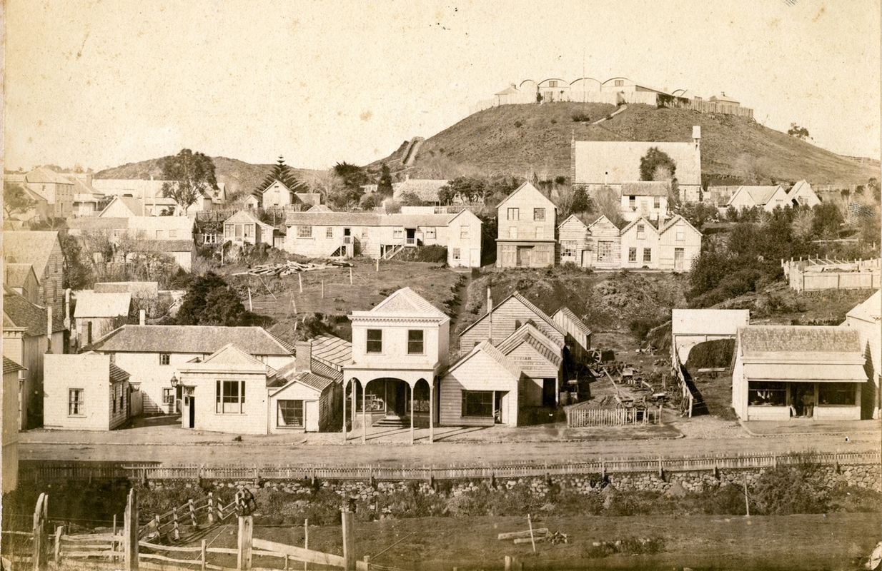 View of 
the barracks on the Marsland Hill, showing the tightly-packed barrel-rooved buildings behind a wooden fance, above the 
buildings among untidy piles of timber in the new town. Some buildings neat, but others ramshackle, and with a large, side-on 
view of the massive church.