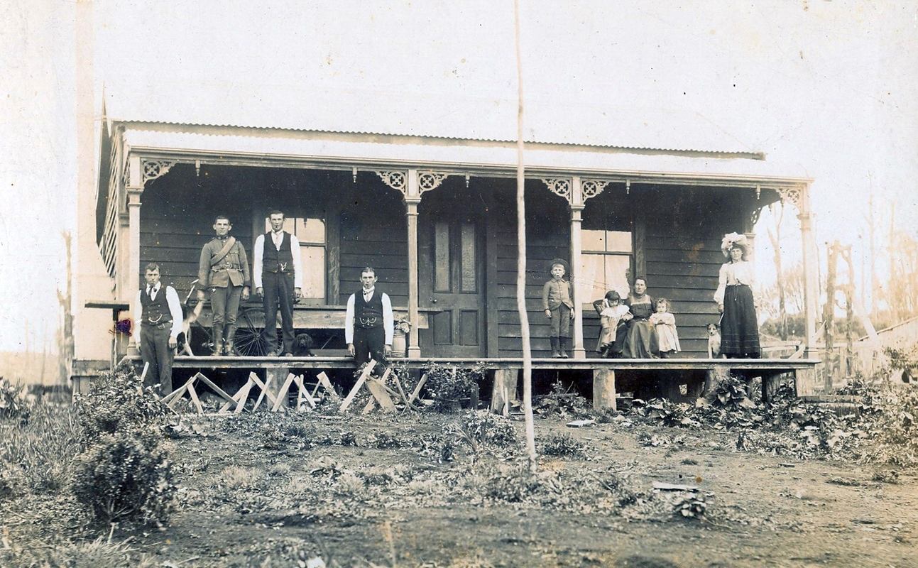 View of the 
front of the farmhouse, showing a front garden of low growth and a few bushes, and some filigree work on the veranda 
uprights