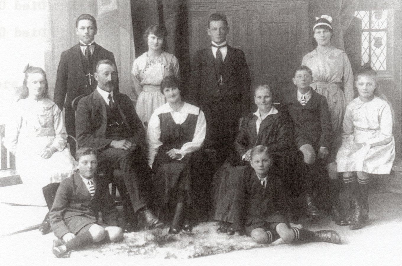 A formal 
family photograph taken inside a formal lounge. in the background is panelling and a leaded window, and on the floor a 
sheepskin rug. August is seated on a chair much higher than his wife. There is quite a gap between Lucy and her mother.