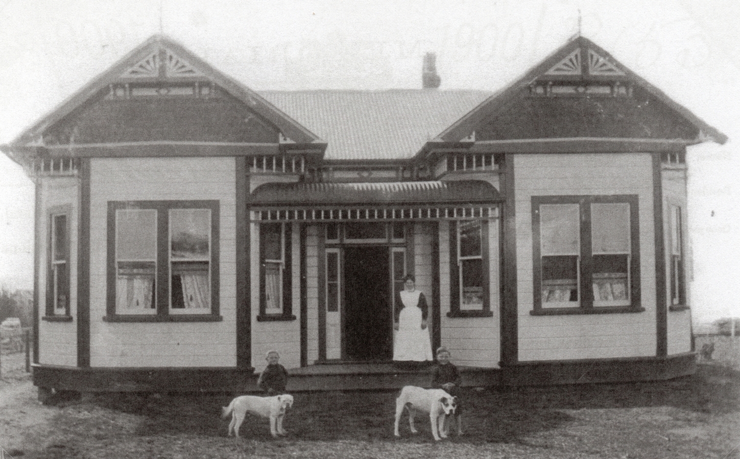 The homestead 
taken from the front. A symmetrical building, and even the two boys behind the two white dogs are on either side of the 
bottom of the steps. a woman in the apron stands to the side of the frint door.