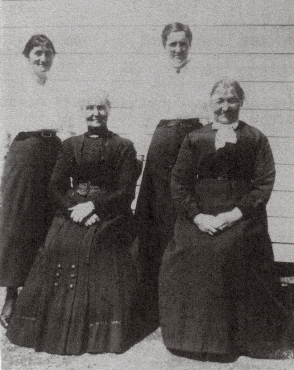 Julia and her sister Anna sitting in front of two young ladies. All are smiiling. Older women have hands on their laps. 
Younger women hands behing them. Older women in dark formal attire. Younger women in dark skirts and white shirts.