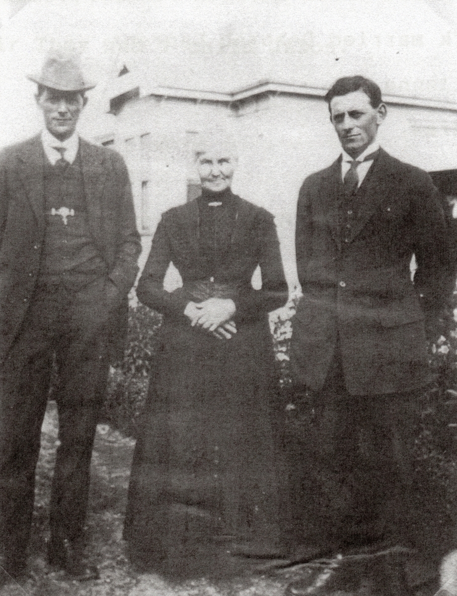 Julia standing 
in the garden between her sons. All smiling but not touching. She has her hands folded in front of her tummy. All in formal 
attire.