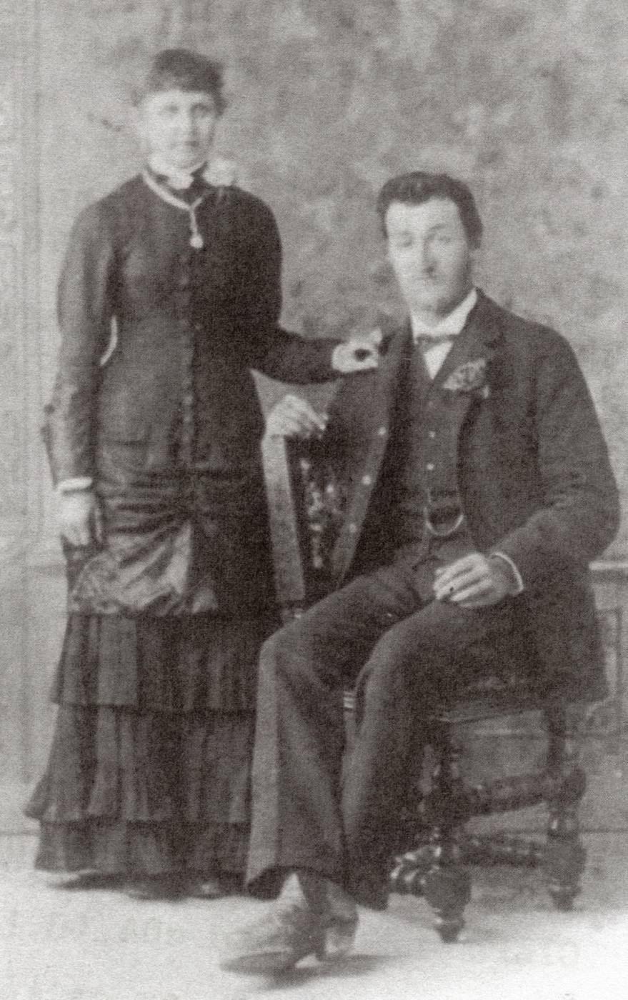 Not an obvious 
wedding photograph. August sitting sideways in a chair and Mary with her hand on his shoulder. Both in formal attire, but 
Mary not in white. Her dress has several flounces and she seems to have a flower on her shoulder and a necklace
