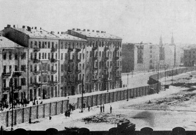 Black and 
white shot of four- and five-storey apartment blocks on one side of the road, the stark wall winding across at an angle, and 
razed emptiness behind the wall. Several people are visible from within the wall.
