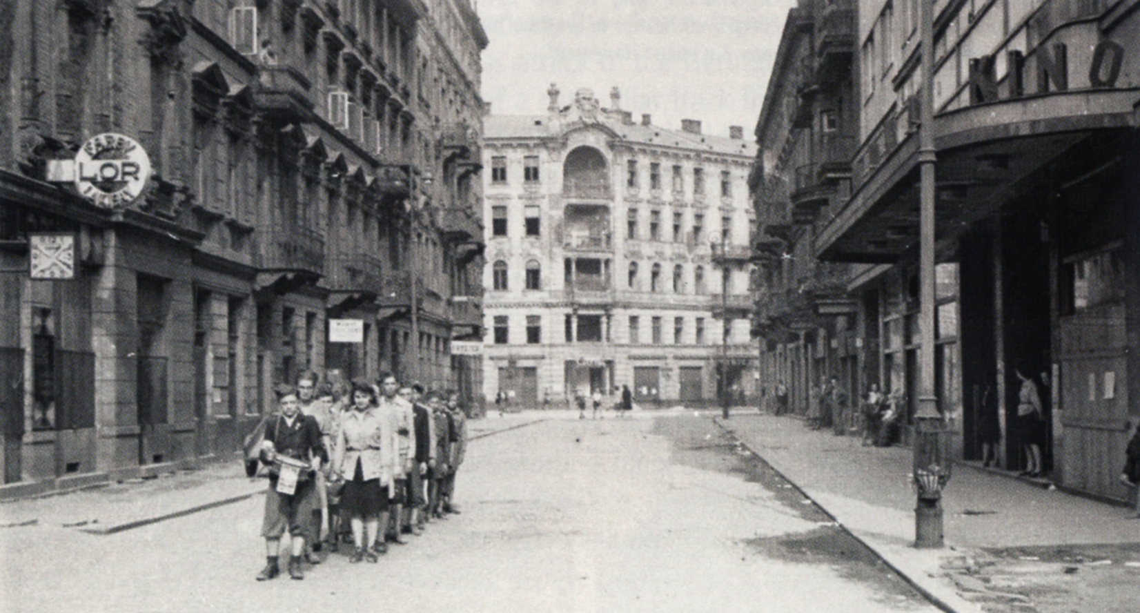 A black and 
white shot of about 20 youngsters in pair formation behind a young drummer, on the left of a road with beautifully facaded 
buildings on either side, and in the background