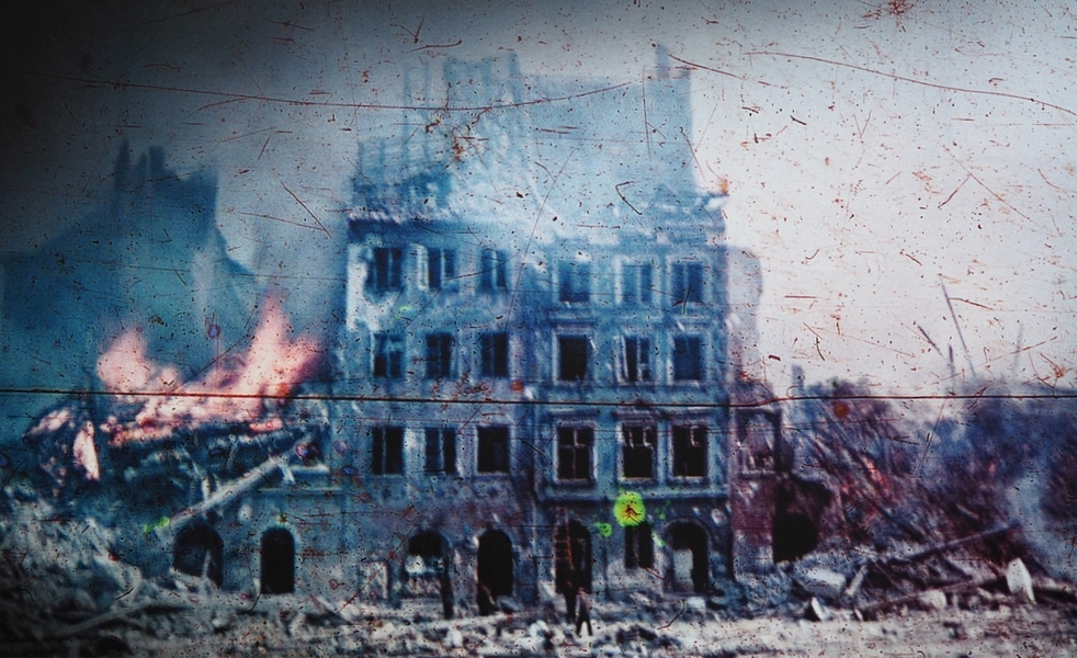 A coloured 
photograph of a building with four storeys still standing, the above destroyed, nothing on one side, and the remains of a 
burning building on the other. Rubble in front.