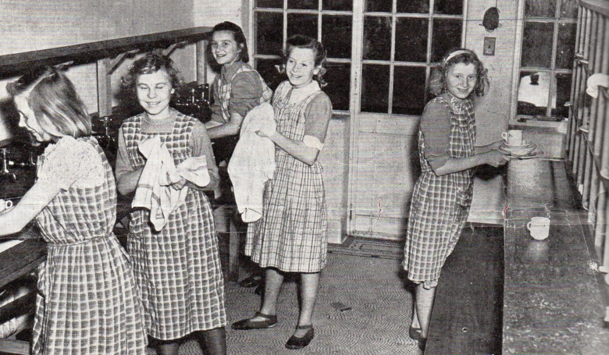 Alina and four others  
drying dishes in their hostel kitchen, all seem happy.