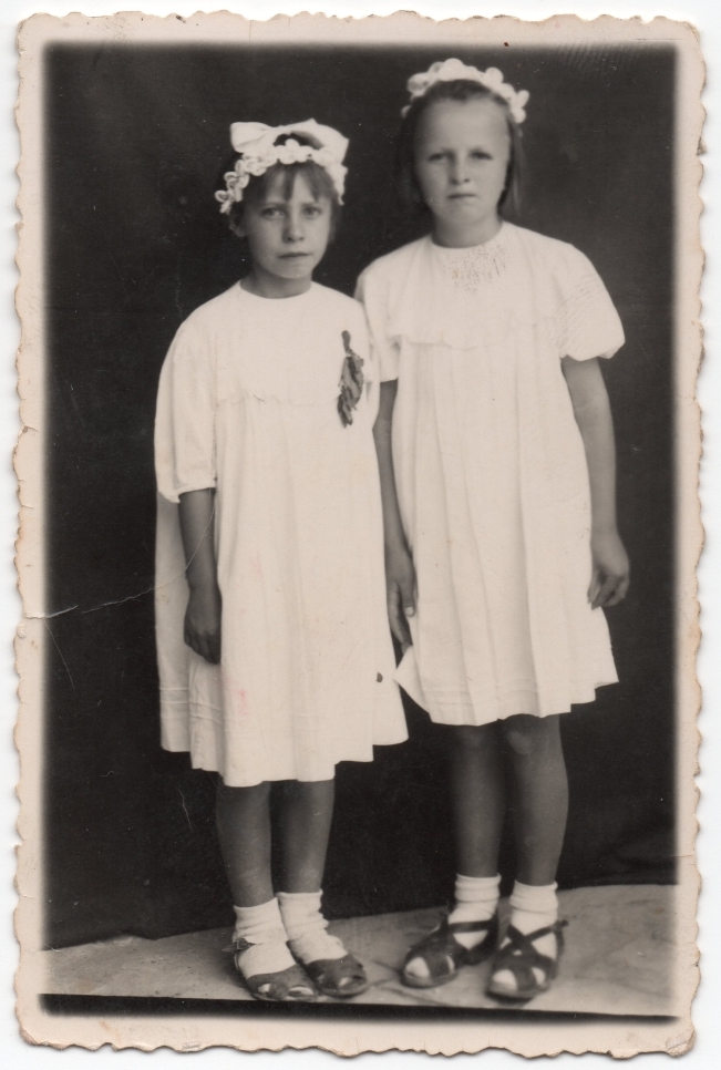 Danuta and a 
friend pose in their white dresses. Plain dark background.