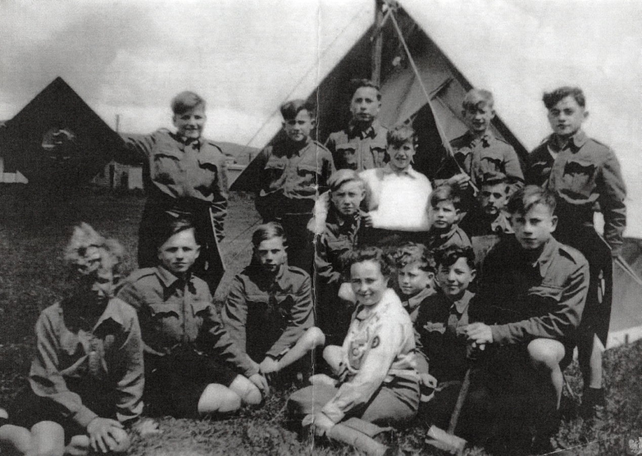 A group of boy scouts 
in front of a tent and with their scout mistress sitting in front.