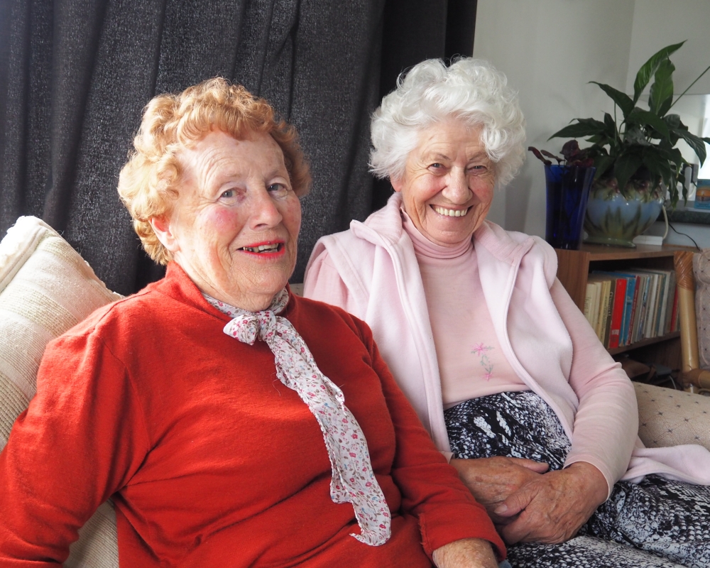 The two old ladies, smiling sweetly, sitting on a couch. Shirley has a red jersey top and a scarf to go with her red hair and Sr Martha is in baby pink jersey to go with her mop of white hair.