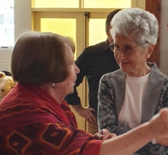 On the right, a taller silver-haired woman is smiling as she listens to a woman in a red-patterened shawl and longer, straight, brown hair.