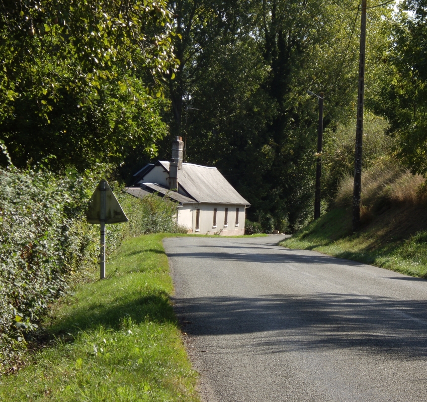 A low-roofed cottage hugging a corner in a road, tall trees in the background.