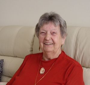 A head and shoulders shot of Janina Iwanica on her couch at home. The couch is cream and she is wearing red and smiling.