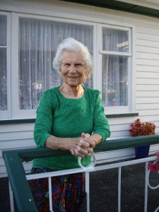 Stasia Kennedy standing outside her house, leaning her arms on the balustrade of her porch. She is wearing an emerald green three-quarter sleeved top over her skirt, and is smiling.