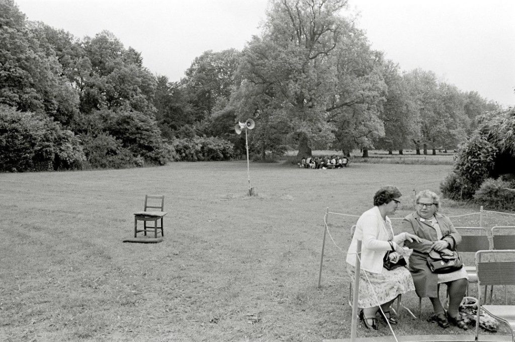 In the right foreground, two old ladies sit on chairs in a roped off area of a large garden. Large trees, including the beginning of a row of the same genus of tree, take up the background. A group sits under the first tree in the row. There is a loudspeaker in the middle and an empty chair to the left foreground, outside and behind the cordon where the ladies are.