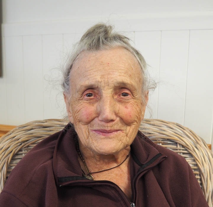 Head and shoulders of a smiling 90-year-old,, hair tied in a bun, but escaping, and in a bit of a brown crumpled track-suit top. Still a handsome woman.

